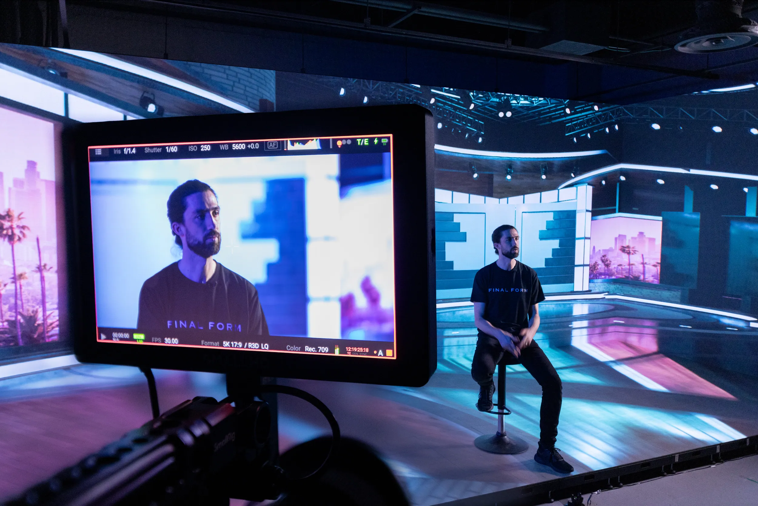 Bearded man in a Final Form tee on a stool in an LED volume showing a neon cityscape, his close-up on a RED camera monitor