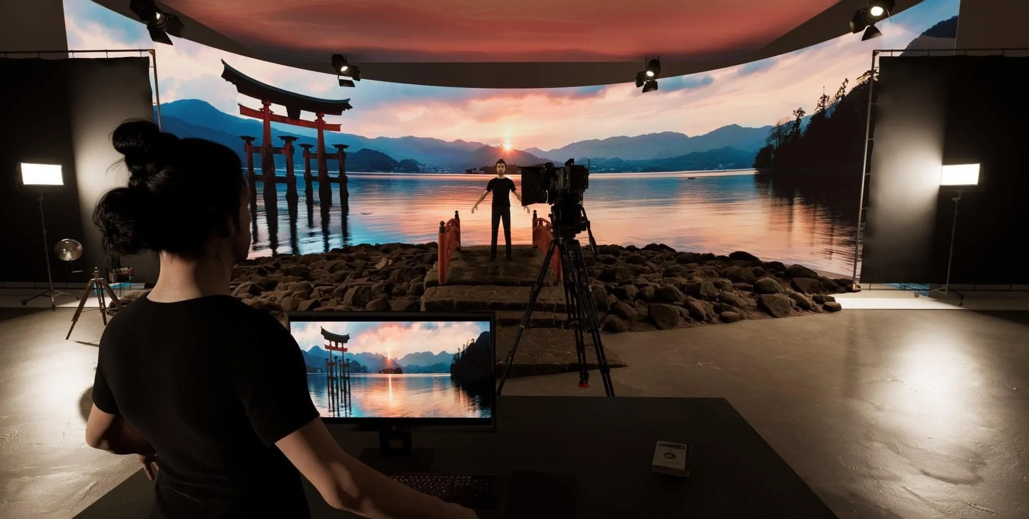 A behind-the-scenes view of a film set depicting a person standing on a rocky pathway near a lake with mountains and a traditional Japanese torii gate in the background. Studio lights and a camera are set up for filming.
