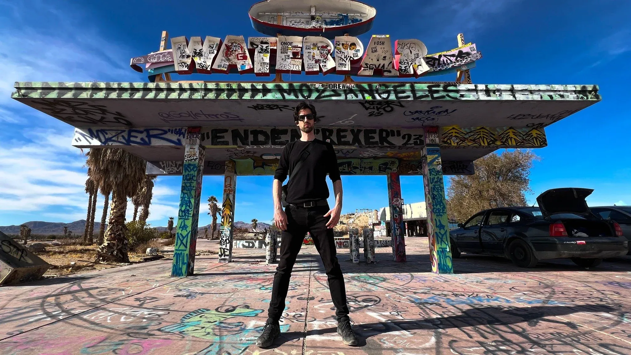 Man in black standing under a graffiti-covered canopy topped with a weathered WATERPARK sign in the desert