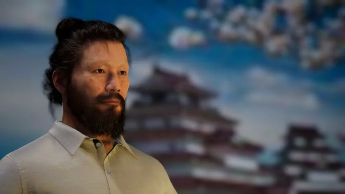 A man with a beard and mustache wearing a beige collared shirt stands in front of a blurred background of a traditional Asian building and cloudy sky.