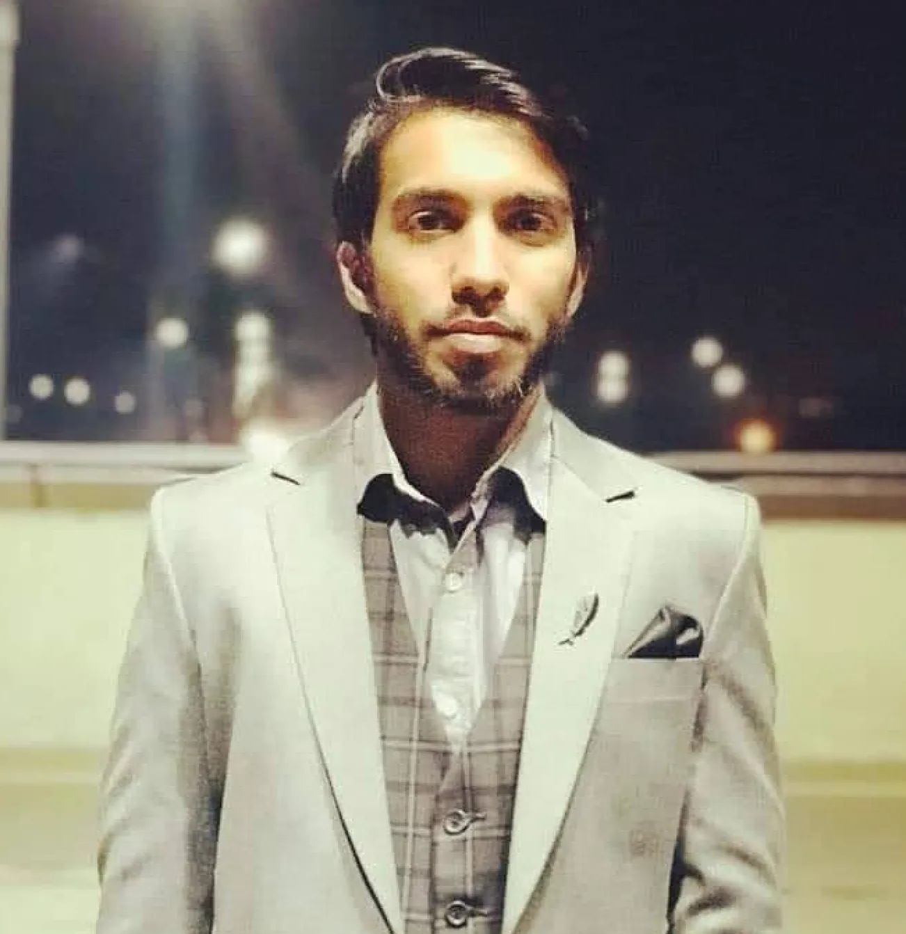 Abdul Arhum in a grey suit and checked waistcoat, photographed at night with blurred city lights behind him