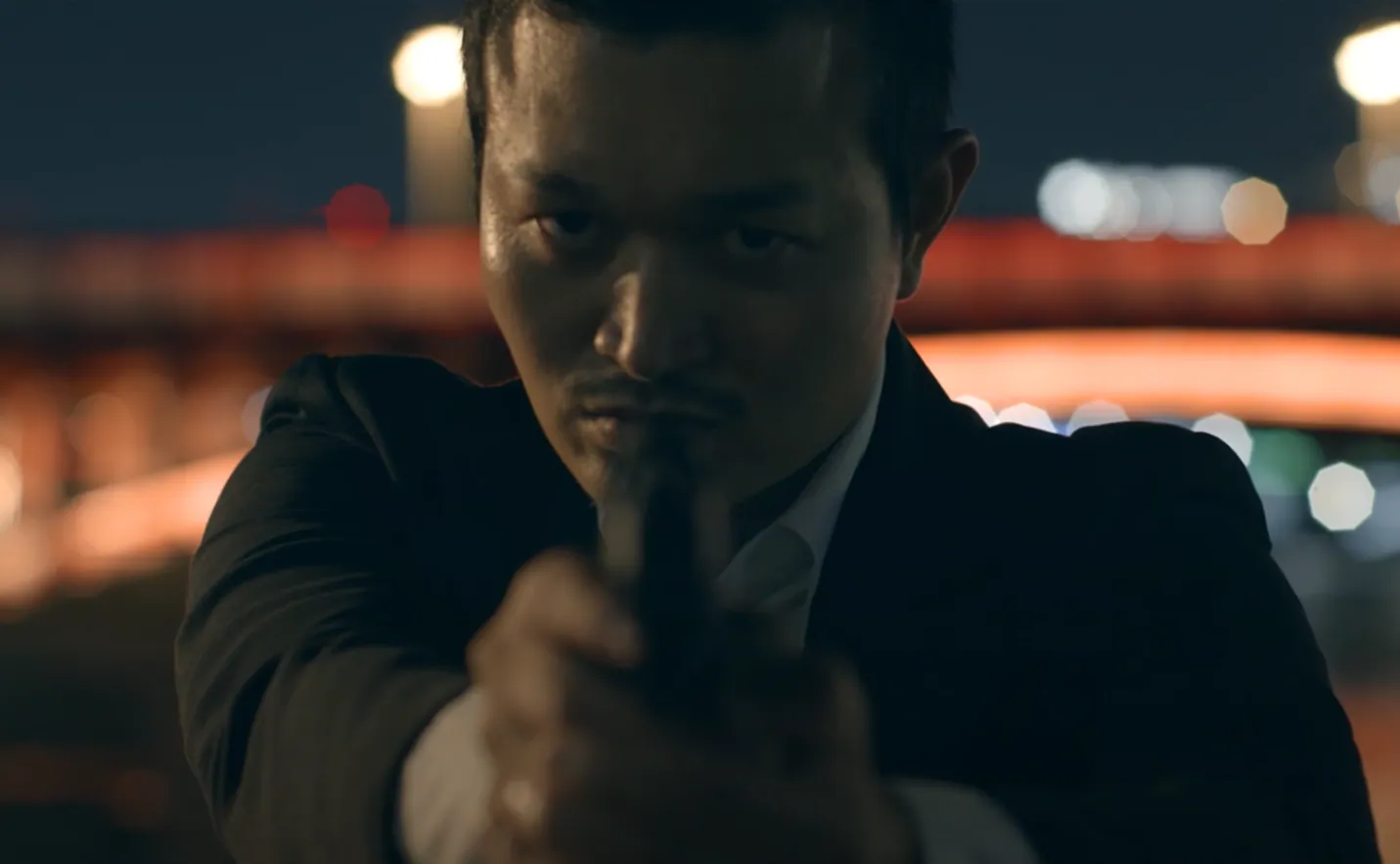 Film still of a man in a dark suit aiming a pistol at the camera at night, a red-lit bridge and city lights blurred behind