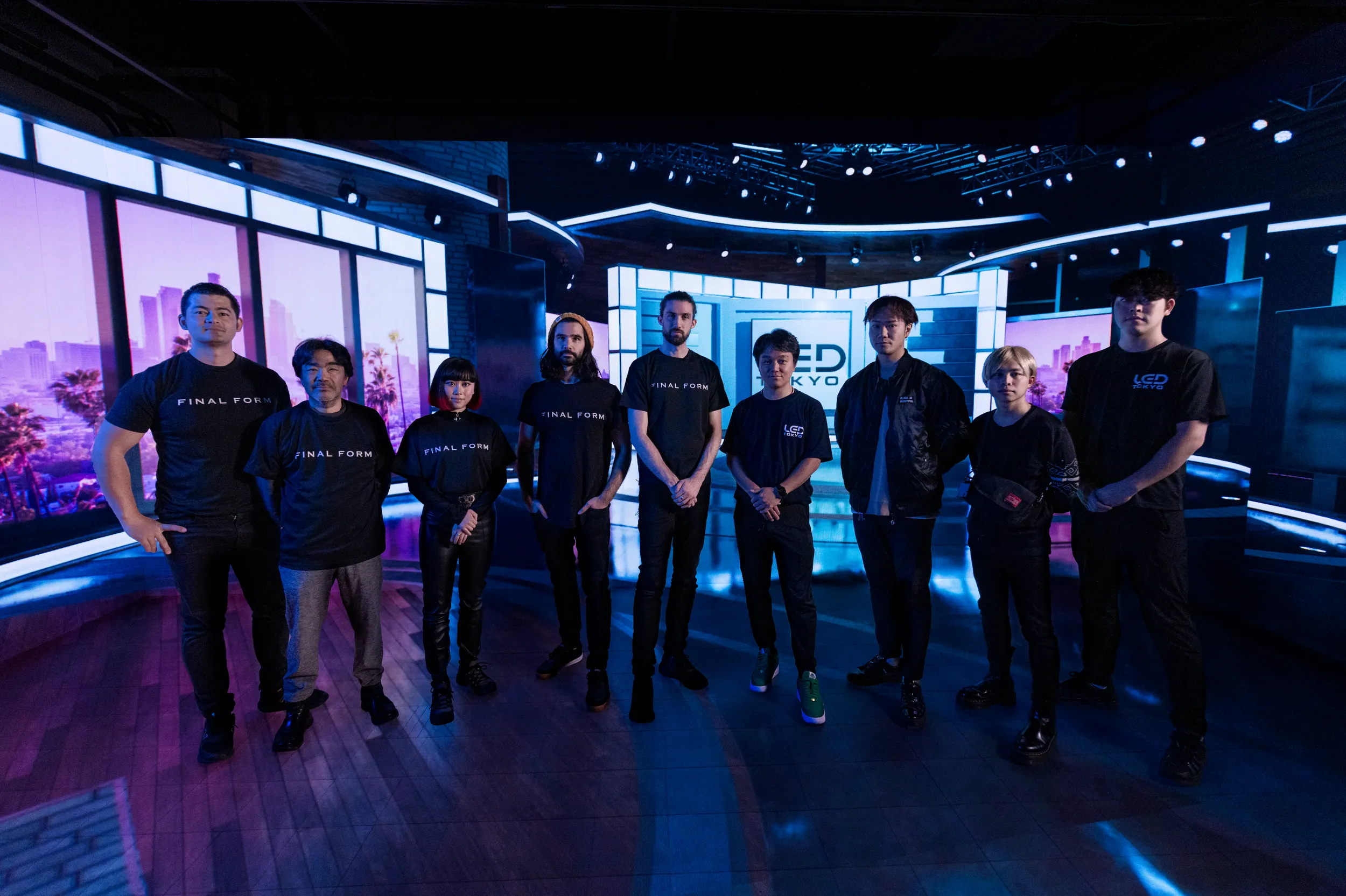 Group of nine people standing on a stage with futuristic lighting, large screens displaying cityscape backgrounds, and a sign that says 'LED TOKYO' in the background.