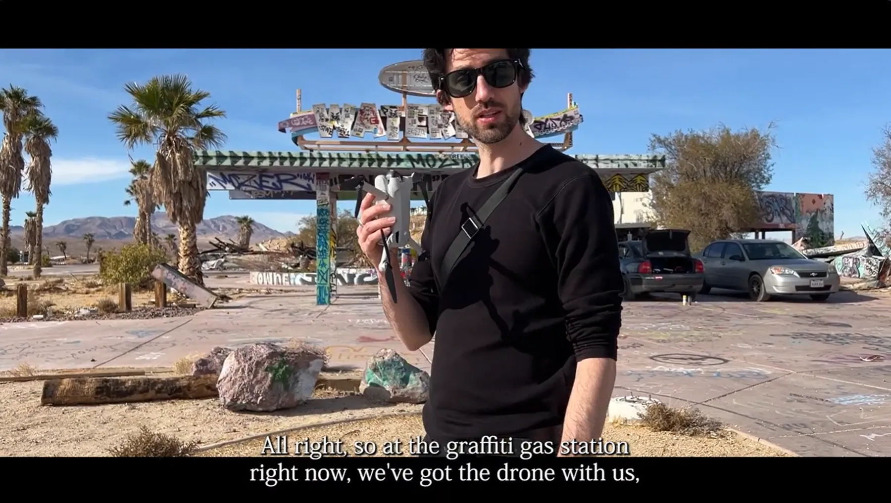 Video still of a man in sunglasses holding a folded DJI Mini 3 Pro drone at a graffiti-covered abandoned gas station