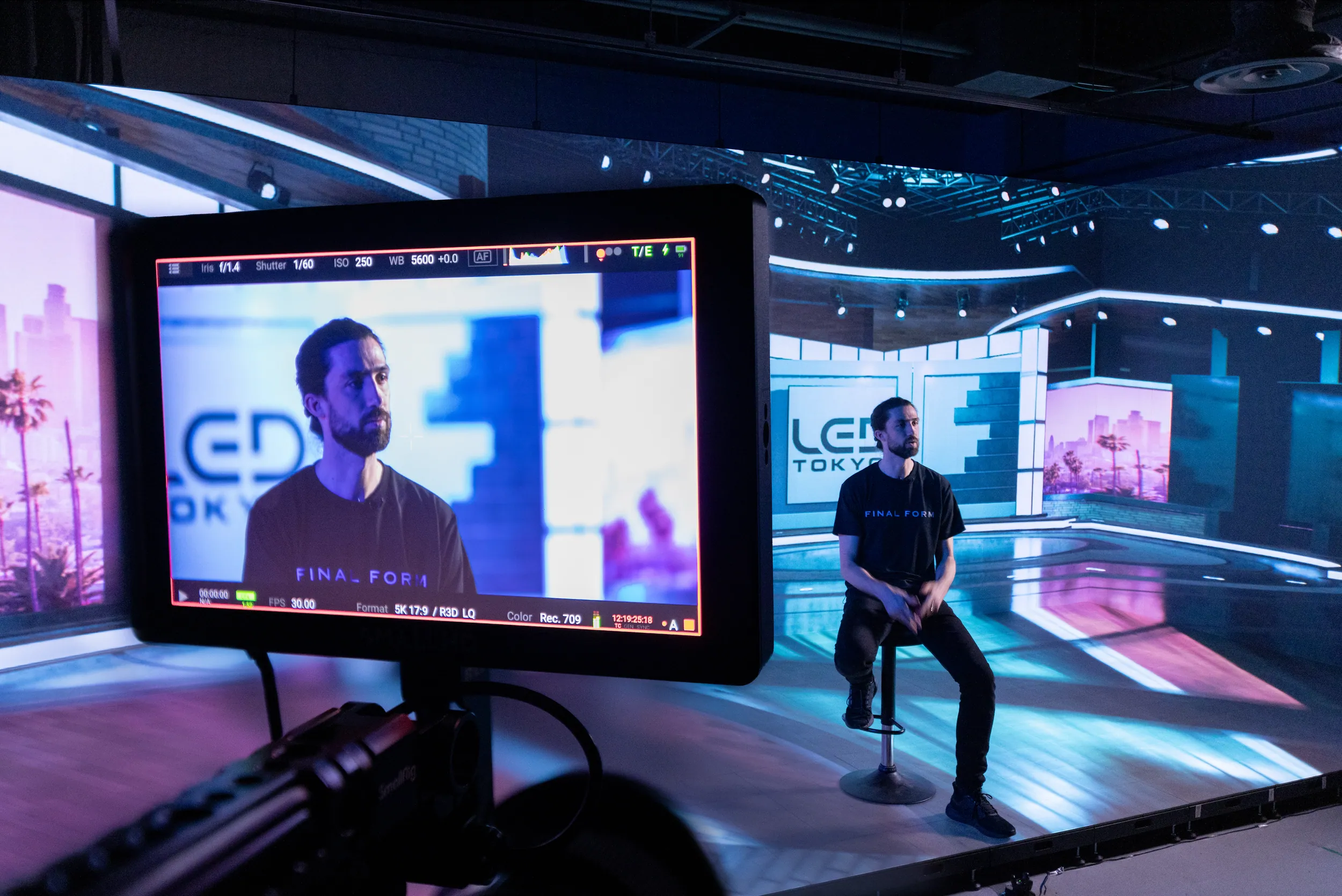 A man sitting on a stool in a television studio with a large screen behind him displaying a stage setup. The camera view is from behind a monitor showing the man on screen, with stage lights and digital backgrounds.