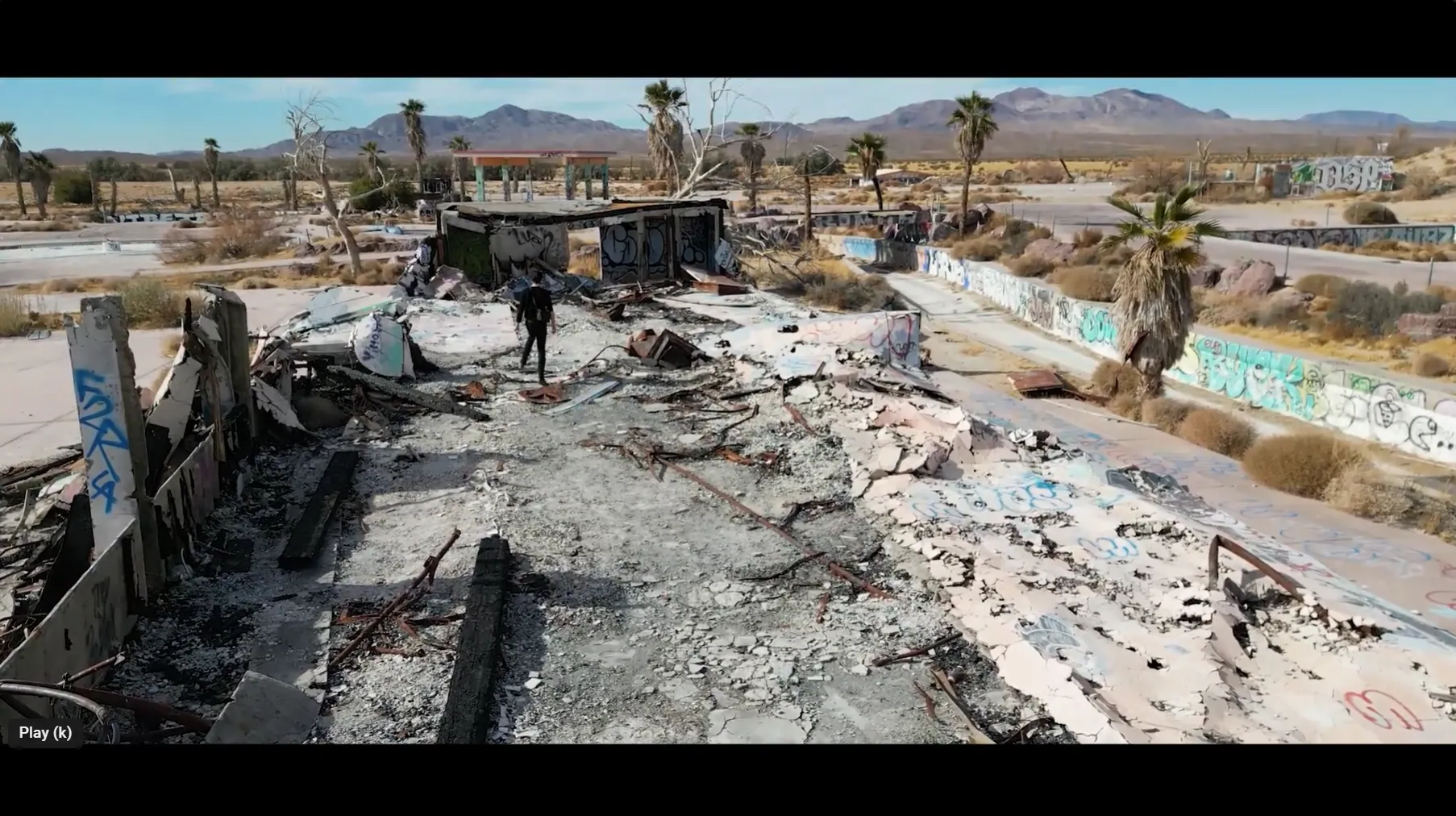 Drone video frame of a lone figure walking through burned rubble and graffiti-covered walls at an abandoned desert park
