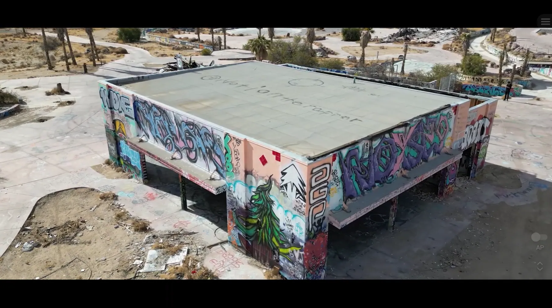 Aerial drone still looking down on a flat-roofed, graffiti-covered concrete pavilion amid palm trees in the desert
