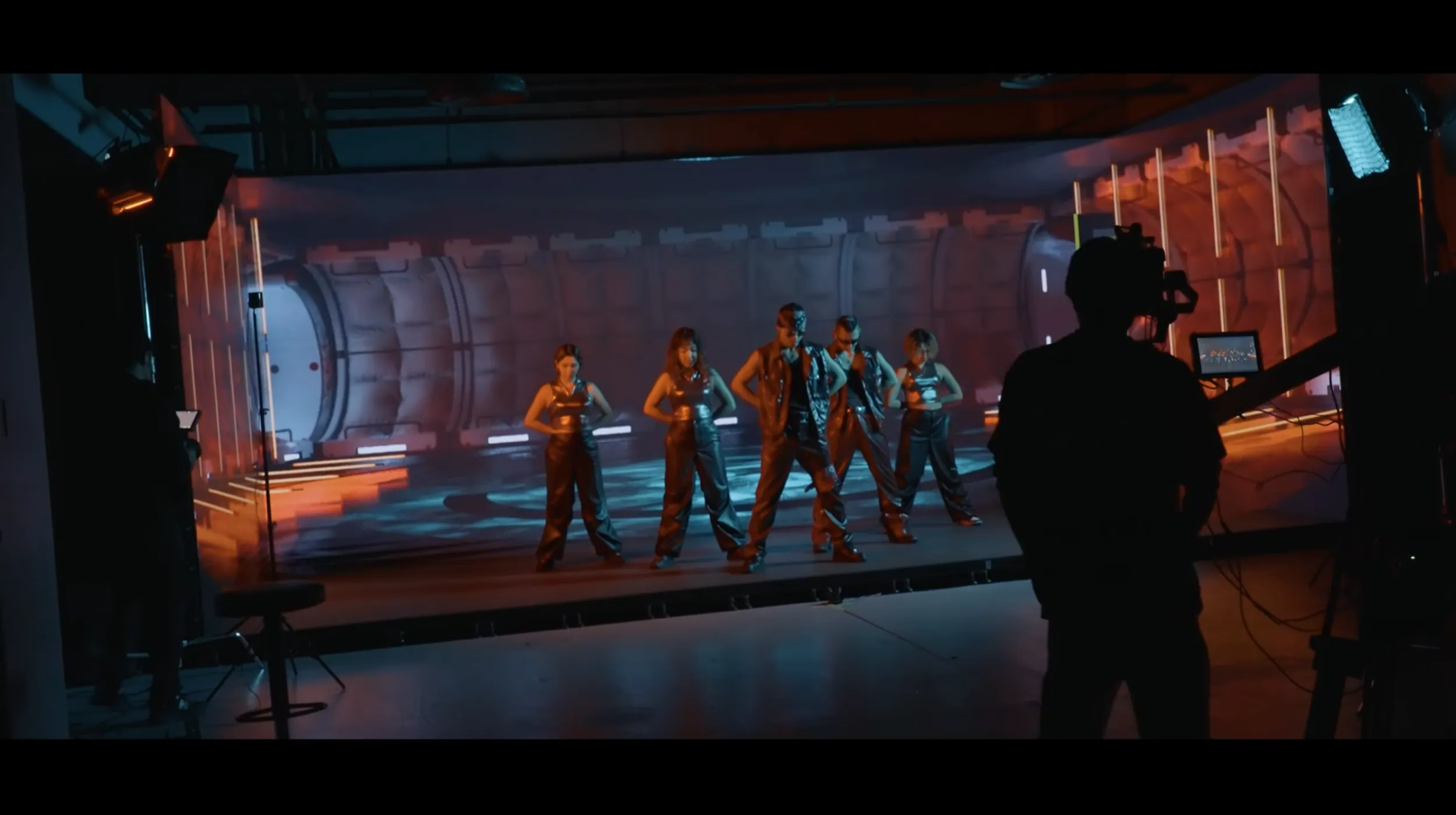Five dancers in silver outfits on an LED stage showing a sci-fi corridor, with a camera operator silhouetted in front.