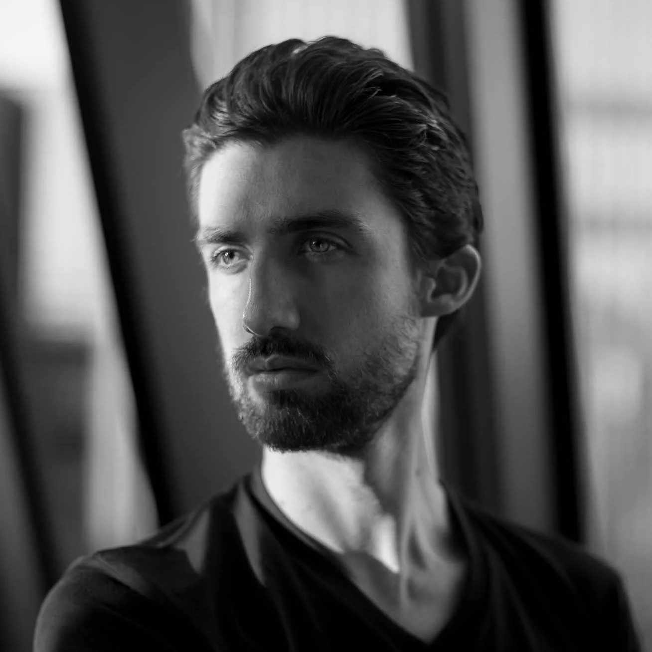 A black and white photo of a young man with dark hair and a beard, looking to the side near a window.