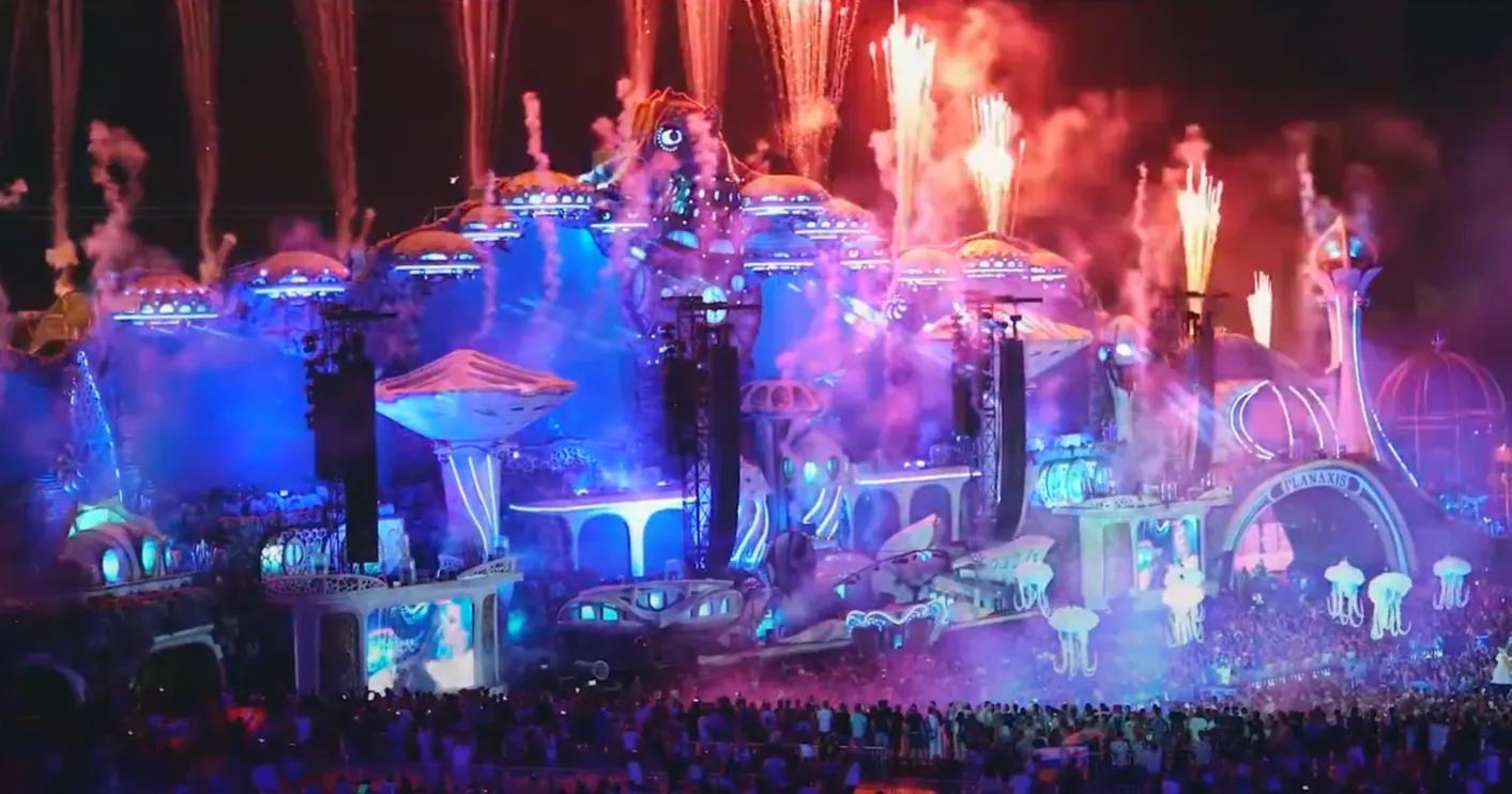 Enormous ocean-themed festival stage at night with fireworks, blue and pink lighting and a vast crowd below.